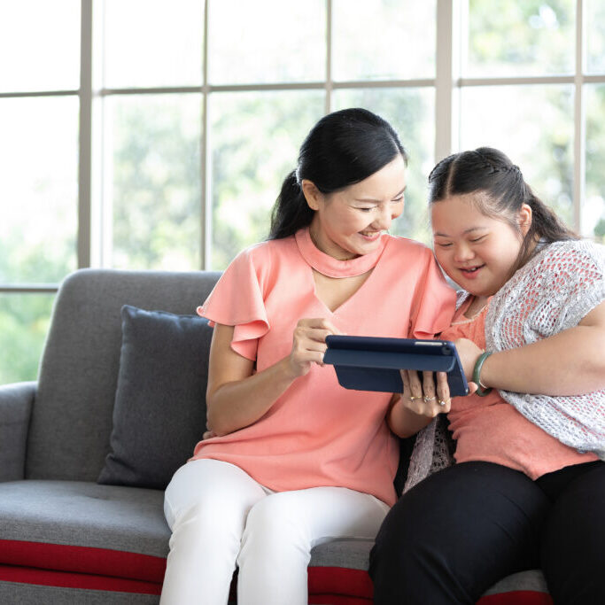 mother using tablet computer with a girl down syndrome or her daughter, smiling and enjoying on sofa